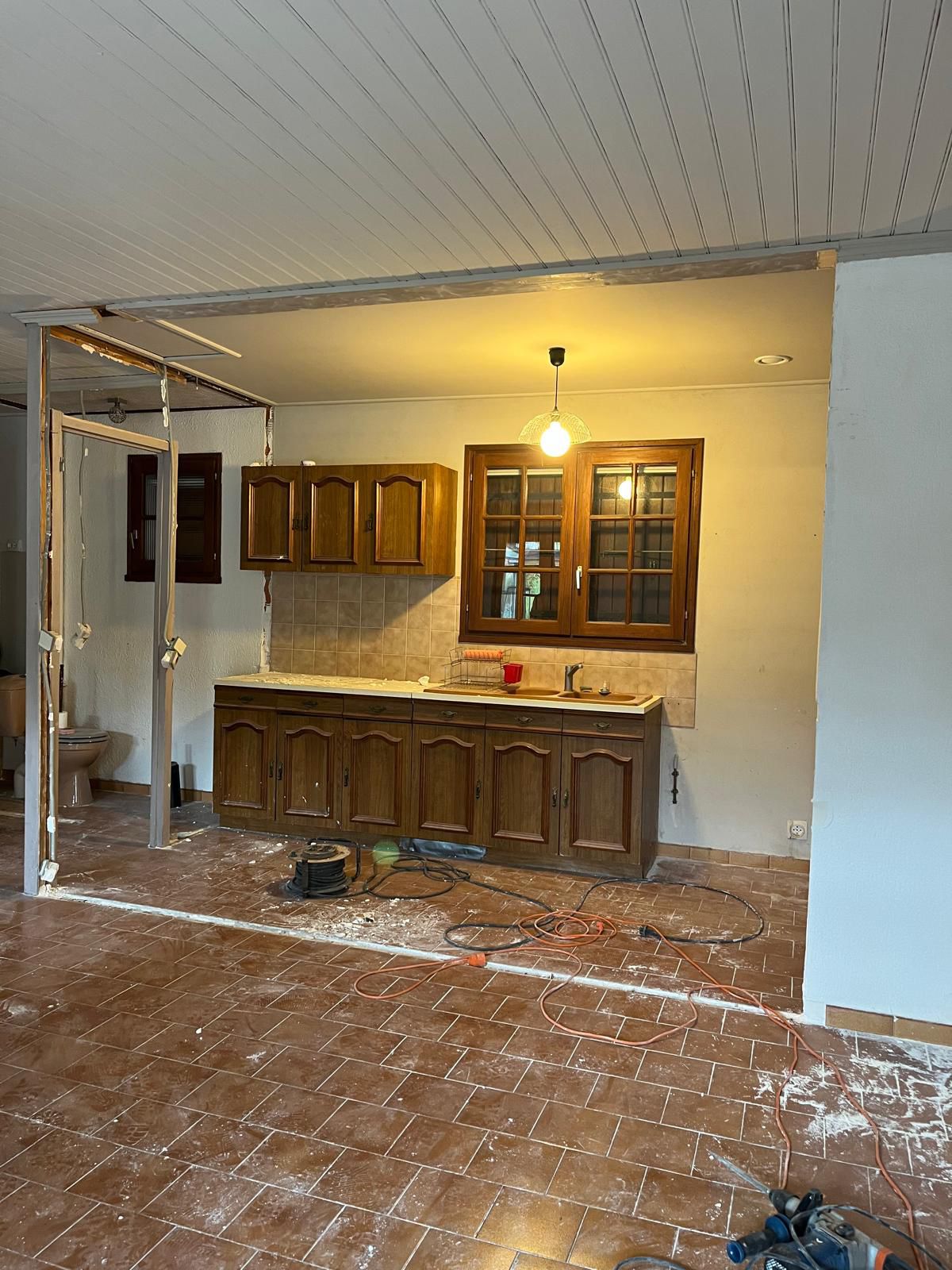 La cuisine avant travaux : meubles en chêne verni, crédence carrelée beige et sol en tomettes, chantier en cours