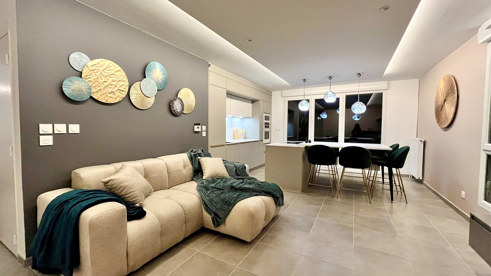 Séjour ouvert aux murs taupe, canapé écru, disques muraux dorés et verts, cuisine et îlot en arrière-plan, Appartement Lumière
