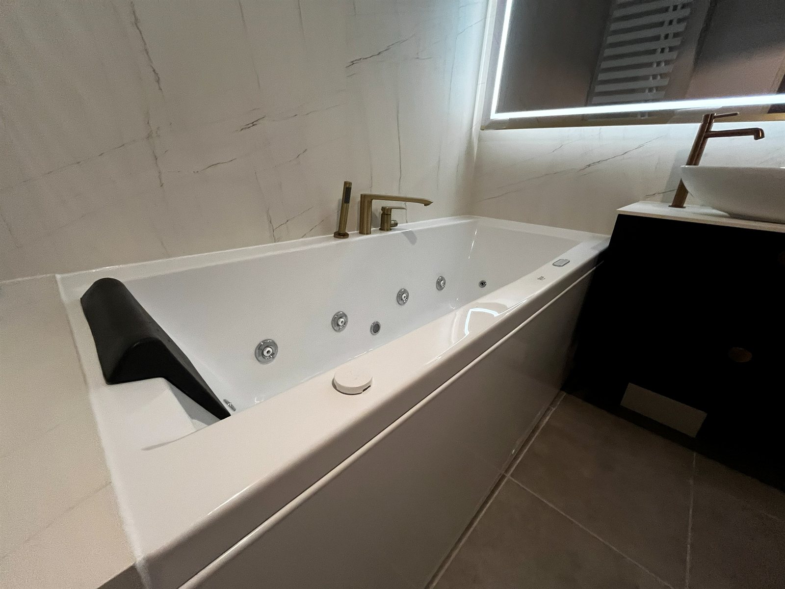 Baignoire balnéo encastrée, murs effet marbre, robinetterie cuivrée et meuble vasque noir