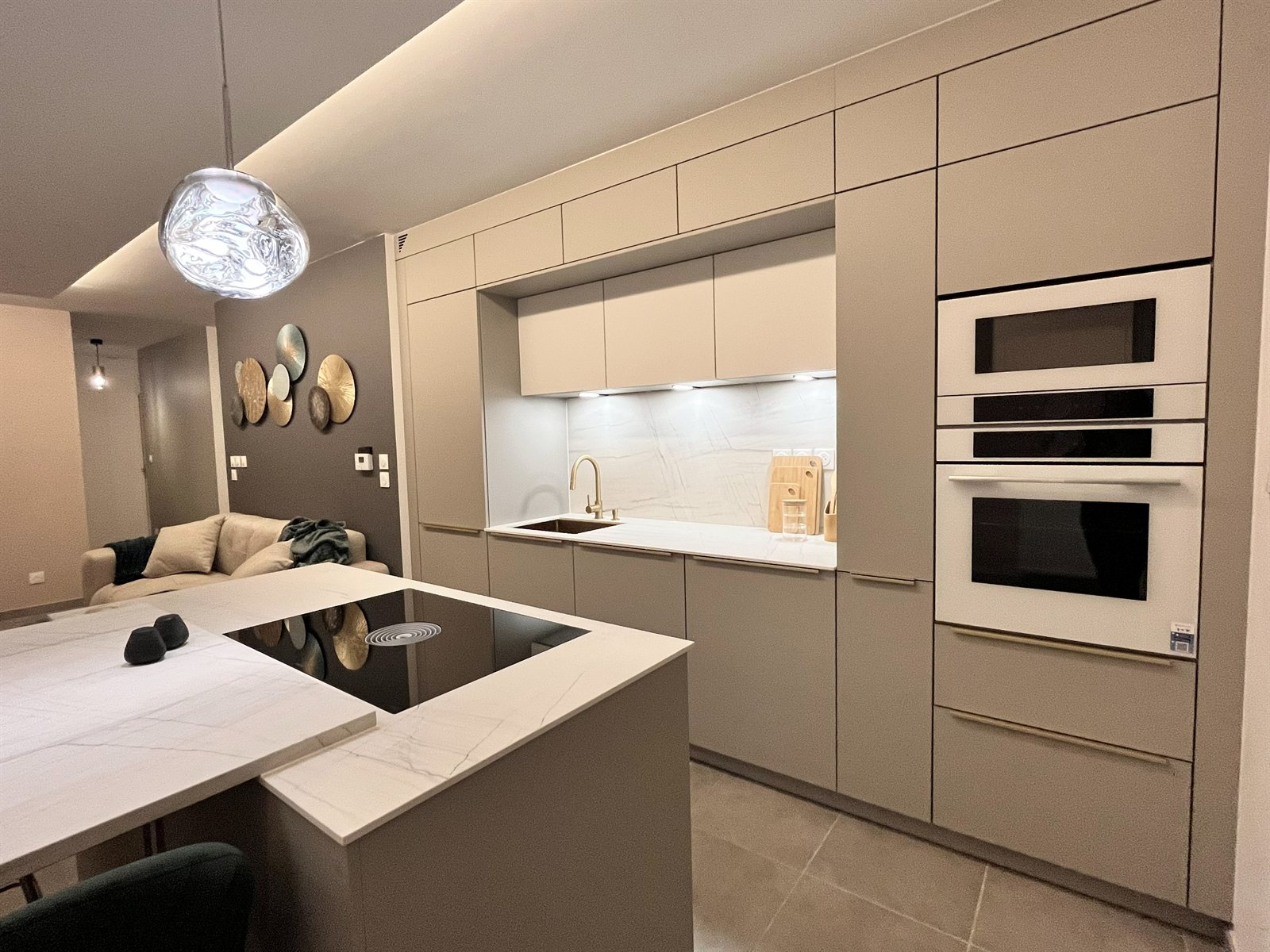 Cuisine linéaire aux façades taupe, fours blancs encastrés, crédence effet marbre et robinetterie laiton